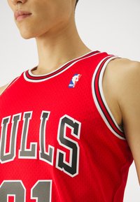 Czerwona koszulka koszykarska z siateczkowej tkaniny, czarno-białe wykończenia, duże napisy "BULLS" oraz logo NBA w górnym lewym rogu.
