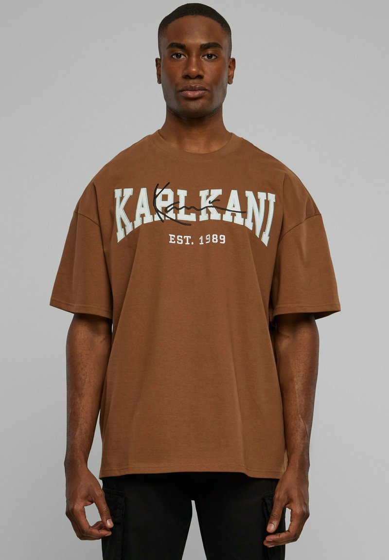 Man die een bruin oversized T-shirt draagt met "KARLKANI EST. 1989" gedrukt op de voorkant, staand tegen een effen grijze achtergrond.