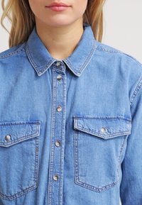 Ljusblå denimskjorta med spetsig krage, knappslutning framtill, två bröstfickor med klaff och tryckknappar, samt en något urtvättad textur.