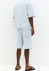 Světle modrá oversize džínová košile s krátkým rukávem v kombinaci s odpovídajícími šortkami délky ke kolenům; má strukturovaný povrch a dvě zadní kapsy.