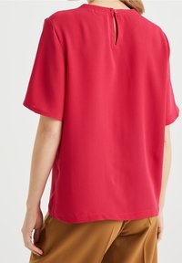 Camicetta rossa a maniche corte con scollatura rotonda e apertura a forma di chiave sul retro, realizzata in tessuto morbido, indossata con pantaloni marroni a vestibilità rilassata.