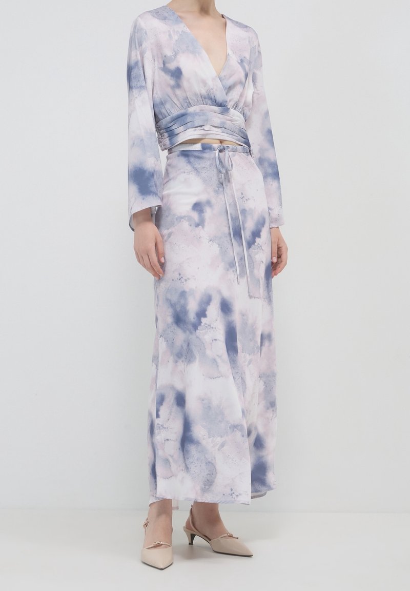 Femme portant un crop top à manches longues et col en V profond, ainsi qu'une jupe maxi taille haute assortie avec un motif bleu et blanc semblable à des nuages.