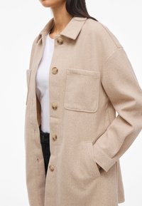 Beige gyapjú keverék dzseki laza fazonnal, kivágott gallérral, két elülső zsebbel és kerek gombokkal. Az anyag puha, textúrált megjelenésű.