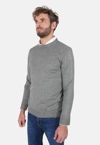 Maglione grigio lavorato a maglia con scollo rotondo e polsini a coste, abbinato a jeans blu. Il tessuto appare morbido e aderente.