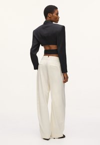 Blazer noir cropped avec épaules rembourrées, taille cintrée et découpe dans le dos, associé à un pantalon large crème et des sandales minimalistes.