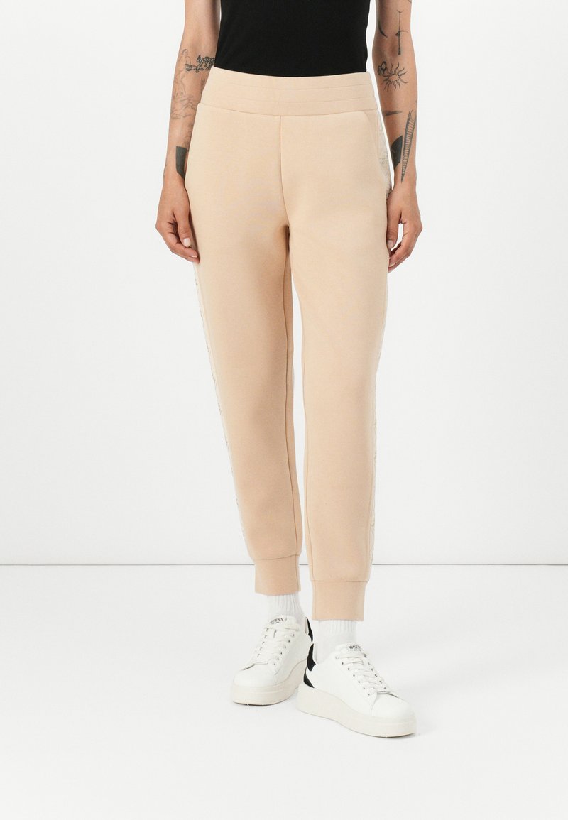 Beige joggers med en slät yta, avsmalnande ankelfållar och sidostrecken. Kombineras med vita sneakers med svarta detaljer.