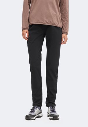 Persoon die een bruin lang mouwen shirt, zwarte broek met rechte pijpen en grijs-zwarte sportschoenen draagt, staand tegen een effen achtergrond.