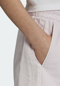 Světle růžové tepláky s bočními kapsami, zdobené vertikálními sklady a měkkou bavlněnou texturou. Šev v světlejší odstínu.