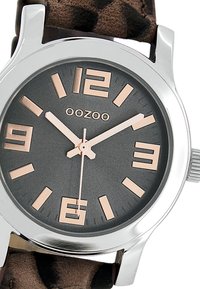 Montre en métal argenté avec un cadran gris foncé, des chiffres en or rose et un bracelet en cuir à imprimé animal brun. Caractérisée par un design minimaliste et des repères clairs.