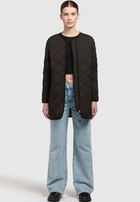 Zwarte gewatteerde lange jas met geometrische patronen, ronde halslijn, ritssluiting aan de voorkant, gecombineerd met lichtblauwe wijde jeans en zwarte schoenen.