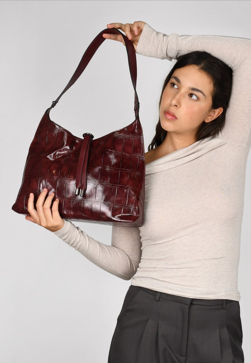 Bolso hobo de cuero burdeos con un patrón de cocodrilo en relieve, base plana y parte superior redondeada. Cuenta con un cierre central de cordón y una correa ajustable.