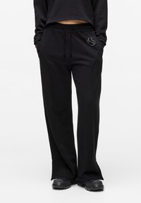 Pantaloni de trening negri cu talie elastică, buzunare laterale și accente decorative din strasuri pe șold. Confecționați dintr-un material moale, cu o croială lejeră.
