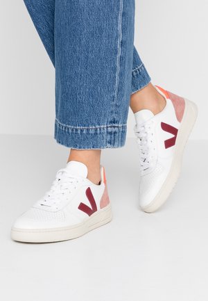 Witte geperforeerde leren sneakers met bordeauxrode en roze suedekleurige accenten, voorzien van een gevoerde zool en veters, tegen een neutrale achtergrond.