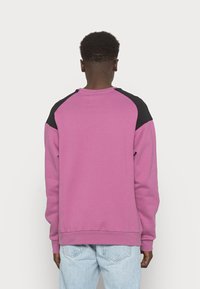 Persona che indossa un felpa rosa con pannelli neri sulle spalle e jeans azzurri chiari, vista da dietro su uno sfondo semplice.