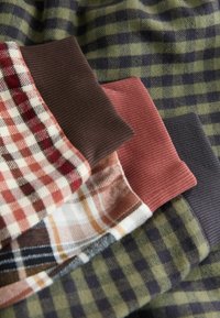 Trīs salocītas auduma gabali ar ribotām aprocēm: viens olīvzaļā un tumši zilā pledā, viens rūsas krāsā ar krēmbalto pledā, un viens brūns ar baltu rūtiņu rakstu.