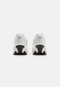 Balti ir kremiški Nike sportbačiai su tekstūruotais paviršiais, apvalia forma, juodu padu ir logotipo detale ant kulno, turintys dėvėjimo kilpą.