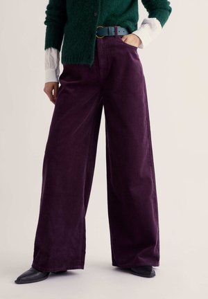 Personne portant un pantalon large en velours côtelé violet foncé, des chaussures noires pointues, un pull vert, une chemise blanche, une main dans la poche.