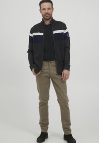 Cardigan cu fermoar de culoare albastru închis, cu dungi albe și bleumarin, tricou negru, pantaloni bej și cizme negre până la gleznă. Textură moale din tricot.