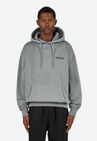 Pelēks kokvilnas kapuci ar priekšējo kabatu, regulējamu kapuci un izšūtu logo. Iezīmēts ar ribotām piedurknēm un apakšmalu, nodrošinot papildu faktūru.