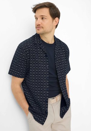 Mann trägt navyblaues, kurzärmeliges Hemd mit Blumenmuster über einem navyblauen T-Shirt, beige Hose und geflochtenem Gürtel, Hände in den Taschen, schaut nach links.