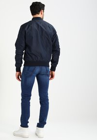 Marine bomberjakke med en glat, skinnende tekstur; ribbet krave og manchetter; parret med blå jeans og hvide sneakers.