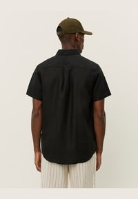 Zwart shirt met korte mouwen, rechte zoom, rug yoke-detail en een kraag. Draag het met licht gestreepte broek en een olijfgroene pet.