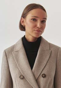 Abrigo de mezcla de lana beige con un diseño de doble botonadura, botones grandes y cuello con muescas, llevado sobre un jersey negro de cuello alto.