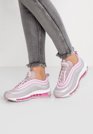 Person iført grå jeans med frynset kant og pinke og grå atletiksko stående på hvidt gulv.