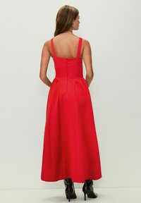 Rotes Maxikleid mit breiten Trägern, tailliertem Oberteil und ausgestelltem Rock. Glatte Textur und Reißverschluss am Rücken. Kombiniert mit schwarzen Stiefeln mit Absatz.