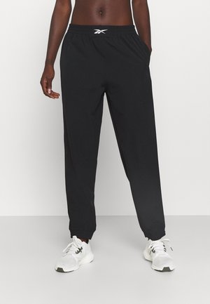 Persoon draagt zwarte joggers met elastische taille en Reebok-logo, staat met één hand in de zak en draagt witte sportschoenen tegen een effen achtergrond.