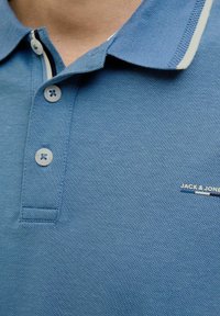 Világoskék pólóing, textúrált anyagból, kontrasztos fehér gallérral és három gombbal. Kis logóval a mellrészen.