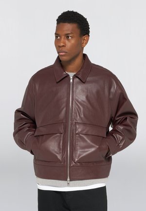 Homme portant une veste en cuir marron foncé avec une fermeture éclair à l'avant et de grandes poches à rabat, debout les mains dans les poches, regardant sur le côté.