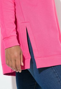 Ruka spočívající vedle růžového dlouhého trička s bočním rozparkem, nošeného s tmavě modrými džínami.