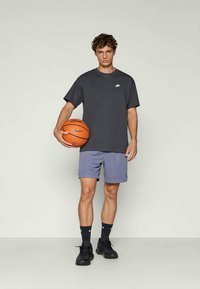 Grijze Nike t-shirt, losse pasvorm, gecombineerd met lichtblauwe sportshorts. Zwarte sneakers en sokken. Houdt een oranje basketbal vast.