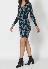 Schwarzes Kleid mit blauem Blumenmuster, langen Ärmeln, V-Ausschnitt, tailliert und knielang. Getragen mit schwarzen Knöchelstiefeln.