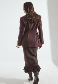 Burgundský šitý blazer so štruktúrovanými ramenami, skombinovaný s textúrovanou hnedou sukňou s strapcovým lemom a s hnedými čižmami po kolená.