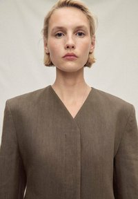 Rjav lana blazer z V-izrezom in strukturiranimi rameni. Gladka tekstura, brez vidnih vzorcev, minimalističen stil.