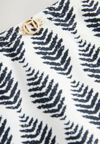 Melnās un baltās krāsas audums ar papardes rakstu, ar apaļu zelta metāla akcentu un krēmkrāsas šuvēm. Mīksta, teksturēta virsma.