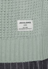 Jasnozielony sweter dzianinowy z ribbanym wykończeniem i wytłoczoną metką z czarnym napisem "JACK & JONES CORE EST. 1990 ENGINEERED DURABILITY."