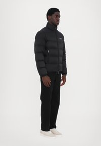 Black puffer jacket with a high collar, zip closure, and a logo on the chest. Worn over dark jeans and light sneakers. Smooth texture.