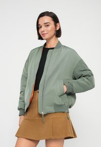 Πράσινο μπουφάν μπόνμερ με πορτοκαλί εσωτερικό, με φερμουάρ, ριμπ μανσέτες και πλαϊνές τσέπες. Φορεμένο πάνω από μαύρο τοπ και καφέ φούστα.