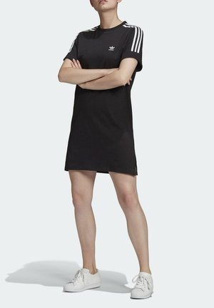 Kobieta stoi ze skrzyżowanymi ramionami, ubrana w krótką czarną sukienkę Adidas z białymi paskami na rękawach i białe trampki na jednolitym tle.