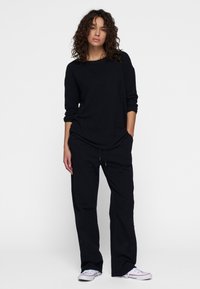 Schwarz langärmliges Oberteil mit weitem Halsausschnitt, kombiniert mit lockeren schwarzen Hosen mit elastischem Bund. Lässige Schuhe runden das Outfit ab.