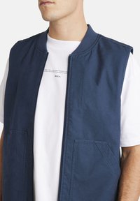 Chaleco sin mangas de color azul marino con cuello acanalado, cierre frontal de cremallera y dos bolsillos laterales. Llevado sobre una camiseta blanca con diseño gráfico.