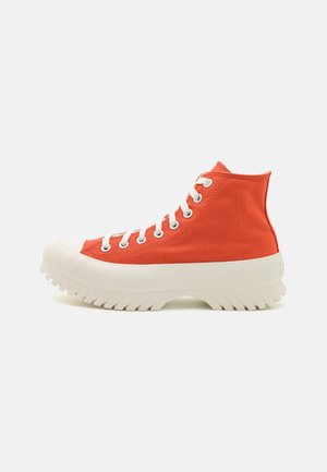 Pomarańczowy sneaker za kostkę z grubą białą, rowkowaną podeszwą i białymi sznurowadłami, widziany z boku na jednolitym tle.
