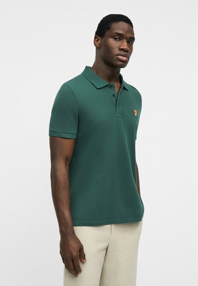 Homme portant un polo à manches courtes vert foncé avec un écusson de renard sur la poitrine et un pantalon beige clair, se tenant devant un fond uni.