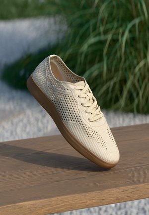 Beżowy casualowy sneaker z ażurowym wzorem i brązową podeszwą, umieszczony na drewnianej powierzchni z rozmytą zielenią w tle.