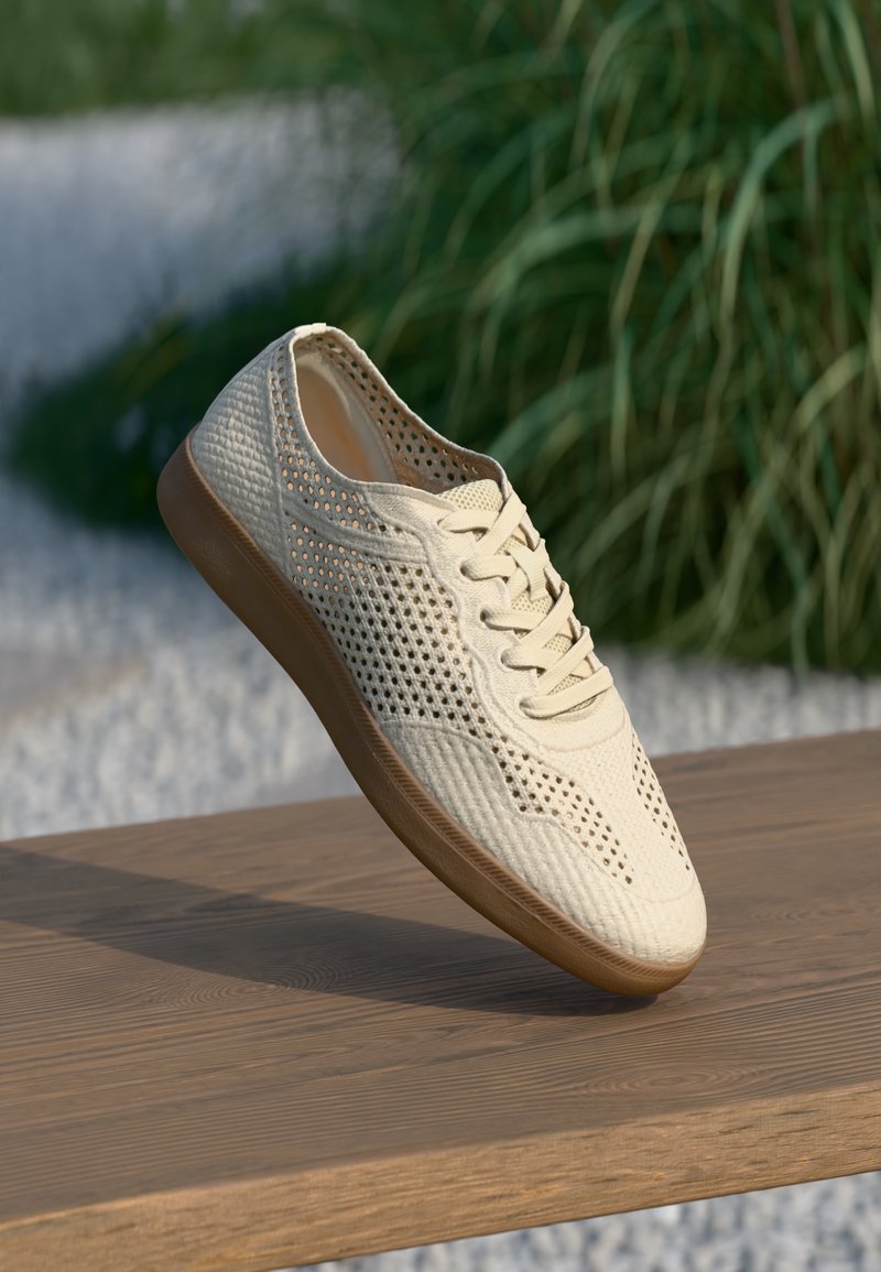 Baskets décontractées beige avec un design perforé et une semelle marron, placées sur une surface en bois avec un arrière-plan flou de verdure.