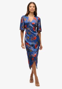 Blauwe bloemjurk met korte pofmouwen, een V-hals, een aansluitende silhouet en een hoge split aan de voorkant tot aan de dij. Draagt heldere sandalen met hak.