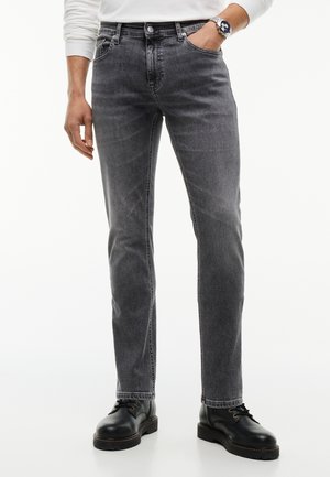 Mann trägt ausgewaschene schwarze Slim-Fit-Jeans, schwarze Leder-Schnürschuhe und ein weißes Langarmhemd mit einer Hand in der Hosentasche.